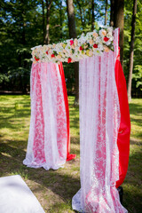 Elegant wedding arch in natural setting. A beautifully decorated wedding arch with white lace and flowers is set up in a lush green forest for a ceremony.