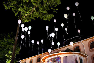 Balloons brighten the festive event. A collection of pastel balloons float gently in the night sky above a lively celebration under bright lights.