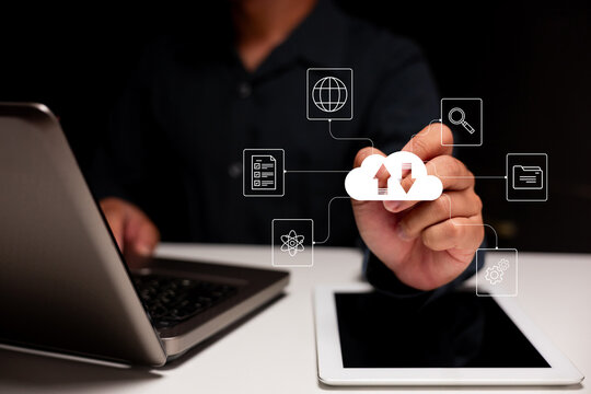A person interacts with digital cloud computing icons representing data transfer and connectivity above a tablet and laptop on a white surface.