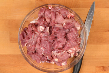A top-down view of a glass bowl filled with diced raw pork, with a knife lying next to it on a...