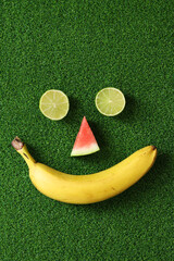 Fruit face on green grass, top view. Two halves of lime, a piece of watermelon and a banana make up...