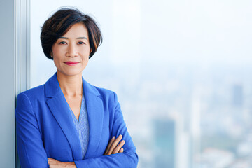 Confident professional woman in a blue blazer stands with folded arms, smiling in a bright office setting.
