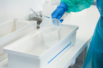Gloved hand carefully pours liquid from calibrated measuring cup into sterile white tray,...