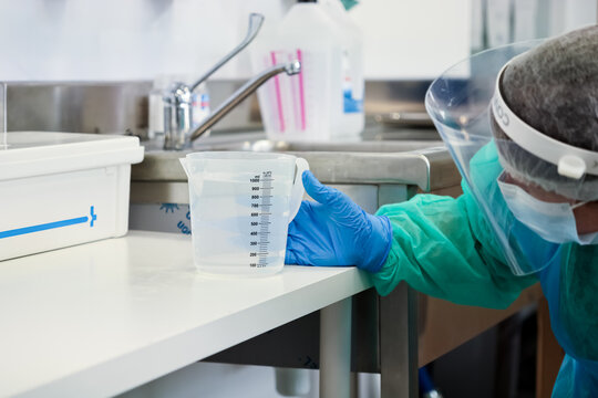 Medical professional in protective gear precisely holding a measuring cup with liquid on a clean laboratory surface, emphasizing safety and accurate scientific measurement