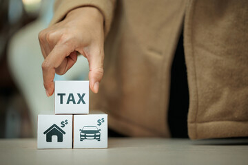 Hand placing wooden block with the word TAX on top of a block with house and car icons, symbolizing...