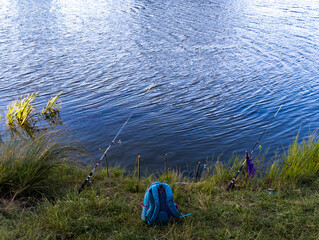 A blue backpack sitting on the shore of a lake next to fishing rods