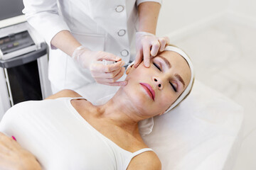 Mature woman getting rejuvenation injection in beauty clinic by cosmetologist in protective gloves and white coat, anti-aging facial treatment to smooth wrinkles and restore youthful skin appearance