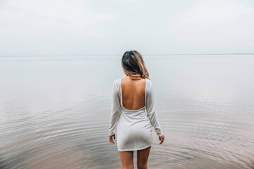 unrecognizable woman standing with back exposed in open white dress near calm lake, facing the water with relaxed posture and serene mood, concept of wellness, spa, fashion editorial