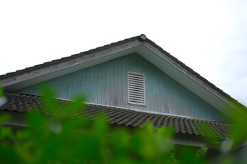 Rustic countryside cottage gable with weathered teal siding, louvered vent, and textured roof tiles, framed by soft-focus green foliage for a tranquil, rural aesthetic.