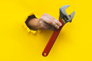 A right hand in a black knitted glove holds a red monkey wrench. Torn hole in yellow paper. The...