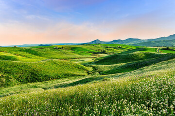 green flowering spring hills in countryside valley with rustic village road among blossoming...
