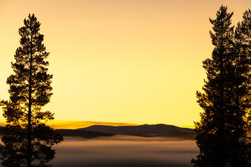 A serene mountain landscape at sunrise, with golden skies illuminating the horizon. A soft blanket of fog drifts over the dark forest below, while the snow-dusted peaks rise in the distance. 