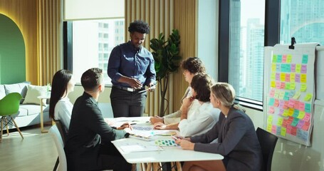 Multicultural team engages in a brainstorming session around a conference table with colorful charts and sticky notes. A man stands to present design ideas.