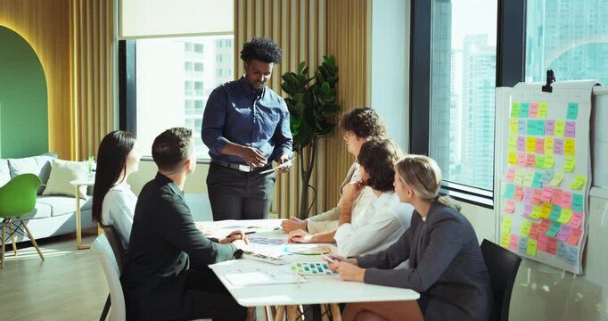 Multicultural team engages in a brainstorming session around a conference table with colorful charts and sticky notes. A man stands to present design ideas. - Powered by Adobe