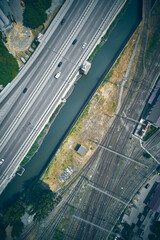 Top-down drone view of multilane highway with moving vehicles, railway yard with numerous tracks, and a narrow canal running between them. Complexity of modern transportation networks