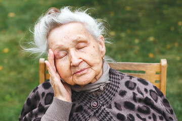 Elderly woman 80-90 years with gray hair with closed eyes touching her head, expressing fatigue or discomfort, sitting outdoors, concept of aging and health awareness