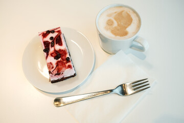 A slice of berry yogurt mousse cake with a chocolate base, served on a white plate next to a cup of cappuccino and a metal fork on a napkin.