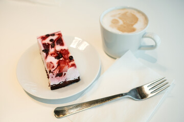 A slice of berry yogurt mousse cake with a chocolate base, served on a white plate next to a cup of cappuccino and a metal fork on a napkin.