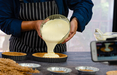 Baker in black and white striped apron pouring creamy custard filling from glass bowl into prepared graham cracker crusts in fluted tart pans kitchen countertop