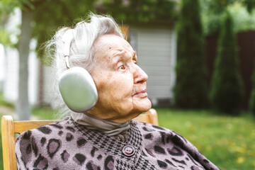 Elderly woman  80-90 years in headphones sitting on chair outdoors and enjoying music, concept of aging and modern technology