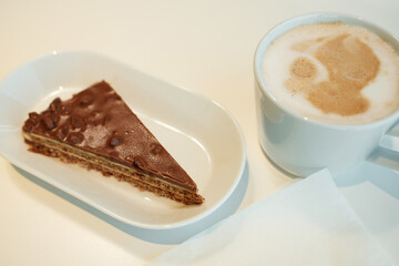 Close-up of chocolate hazelnut cake slice with layers and crunchy topping next to a frothy cappuccino.