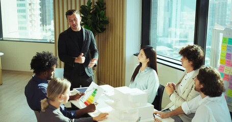 Multiracial group discussing ideas around table in bright office space, reviewing charts and architectural models during a team meeting.