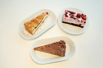 Top-down shot of three different cakes on white plates: berry cheesecake, chocolate hazelnut slice,...