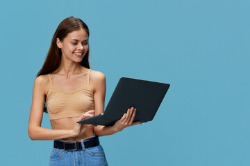 Young woman holding laptop with smile, wearing casual beige top and blue jeans, standing against...