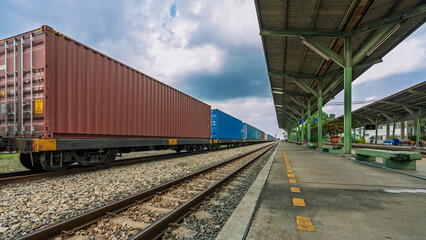 Obraz premium Freight train with container cargo standing at rural railway station platform under cloudy sky, transportation and logistics concept for supply chain, cargo delivery, and intermodal shipping industry