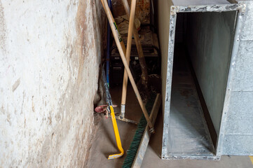 Brooms, shovels, and a metal box are organized against a rough wall in a construction space,...