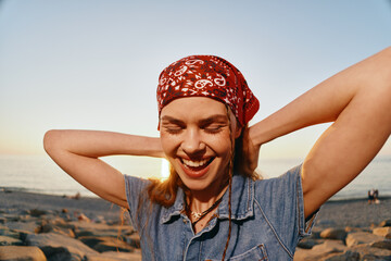 smiling woman with red bandana enjoying sunset on rocky beach in casual denim vest and natural warm...