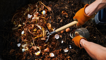 Hands with orange gloves using a garden fork to turn compost in a dark bin filled with organic materials, showcasing sustainable waste management practices and eco-friendly gardening