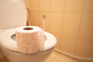A roll of toilet paper sits on the rim of an empty modern toilet in a clean, tiled bathroom. Light cream tiles emphasize the simple decor and practical utility of the space