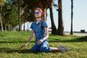 young woman sitting on green grass wearing blue denim outfit and red headscarf outdoors in sunny...