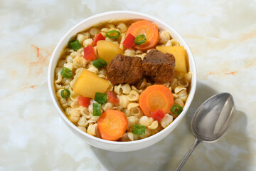 Hearty beef and vegetable soup with noodles, in a white bowl on a marble base.