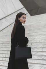 Designer luxury handbag featured with model in monochrome outfit on marble modernist building stairs, showcasing urban architecture and contemporary fashion branding with cinematic composition.