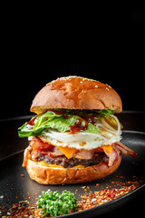 Gourmet burger with bacon, egg, jalapenos, cheese, lettuce, onions, and barbecue sauce on a sesame bun, served on a black plate with herbs and seasoning.