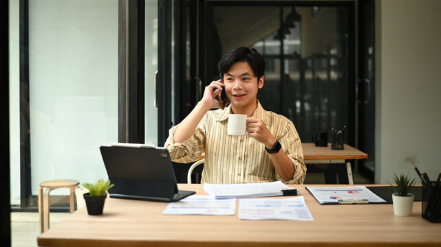 A Young Asian man have a conversation during a coffee break in a workplace setting, with financial reports on the table - Powered by Adobe