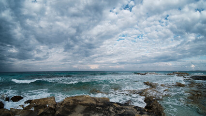 Wide-angle view of a rocky seashore with waves crashing against rocks under dramatic, cloud-filled sky. Raw power of nature, dynamic interaction between sea and sky