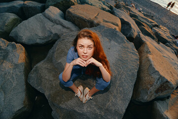 young woman sitting on large rocks by the sea wearing casual blue outfit and sandals during golden...