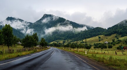 Fototapeta premium The road leads to the mountains, with tree-lined mountain roads, cloud-shrouded mountains in front of and behind, and damp asphalt pavement. Forest after rain with misty mountain background.