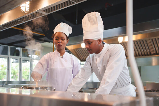 Team of African American man and woman chefs working together in modern kitchen, wearing white uniforms and hats, checking food in steaming pot.