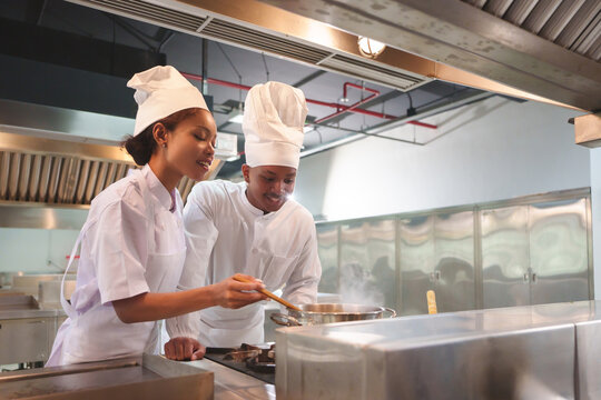 Team of Black male and African American female chefs in white uniforms cooking together, woman stirring food, teamwork in kitchen.