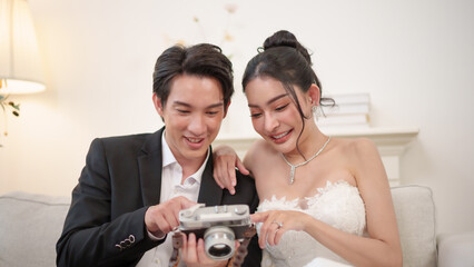 Happy Asian Bride and groom cuddle on sofa, looking at their wedding photos on camera. A calm and...