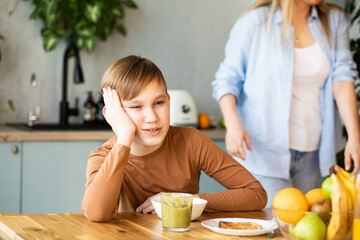 Kid boy teenager with poor appetite sitting the table in kitchen at home and doesn't want to eat.