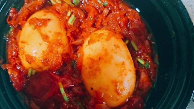 ​A close-up of two whole, peeled hard-boiled eggs nestled in a rich, vibrant red sauce that appears to be spicy and tomato-based, possibly a curry or stew.
