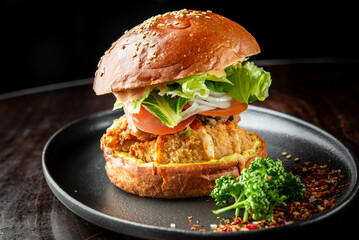 Close-up of a gourmet fried chicken sandwich with lettuce, tomato, onion, and sauce on a sesame seed bun, served on a black plate, garnished with parsley and chili flakes.