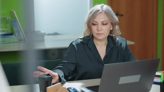Accountant takes quick break to sip hot latte before continuing typing on laptop, working at modern office desk with natural light, documents, and relaxed but professional demeanor