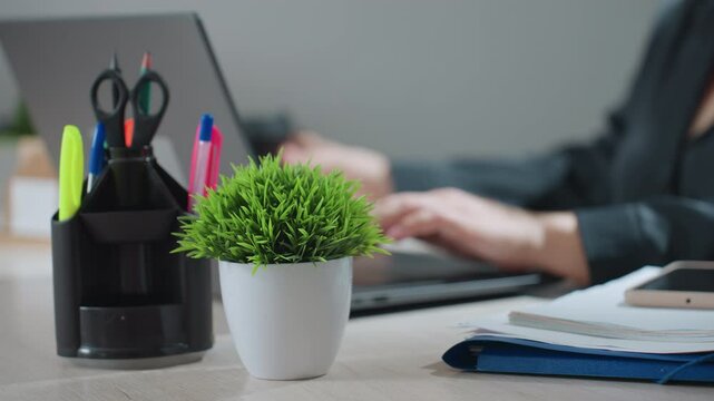 Close up of woman typing on laptop in bright workspace, pauses to reach for coffee then drops hand and continues working, surrounded by colorful office supplies and indoor plant