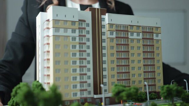 Close up of professional woman dressed in black carrying detailed building model toward office desk, showcasing modern real estate concept and architectural miniature with urban layouts